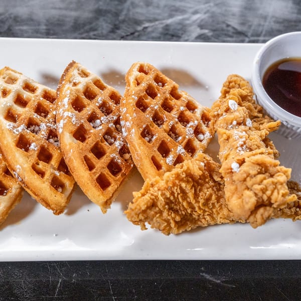 Golden waffles with powdered sugar and crispy fried chicken tenders with syrup on a white plate.