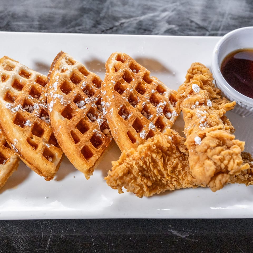 Golden waffles with powdered sugar and crispy fried chicken tenders with syrup on a white plate.