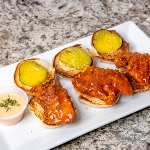 Spicy chicken sliders with pickles and a side of dipping sauce on a white plate.