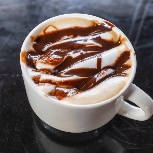 Close-up of a white mug filled with a frothy drink, drizzled with chocolate syrup, on a dark surface.