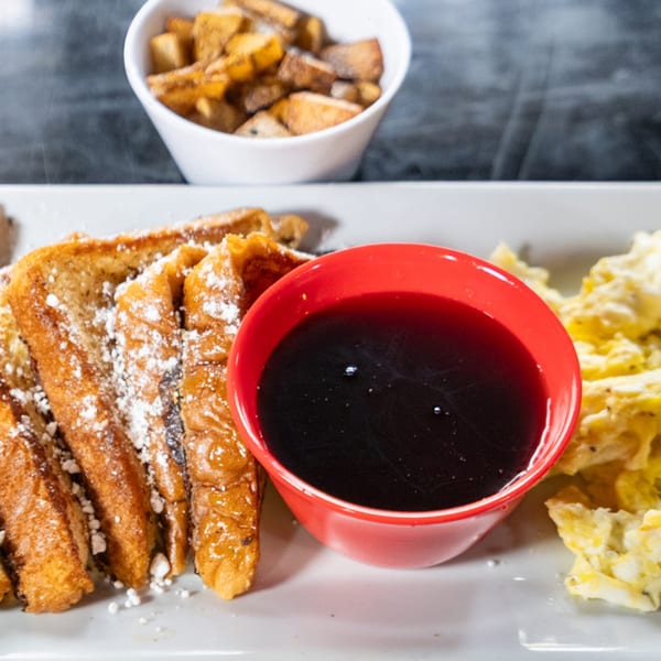 French toast with powdered sugar, a red bowl of syrup, scrambled eggs, and a side of home fries.