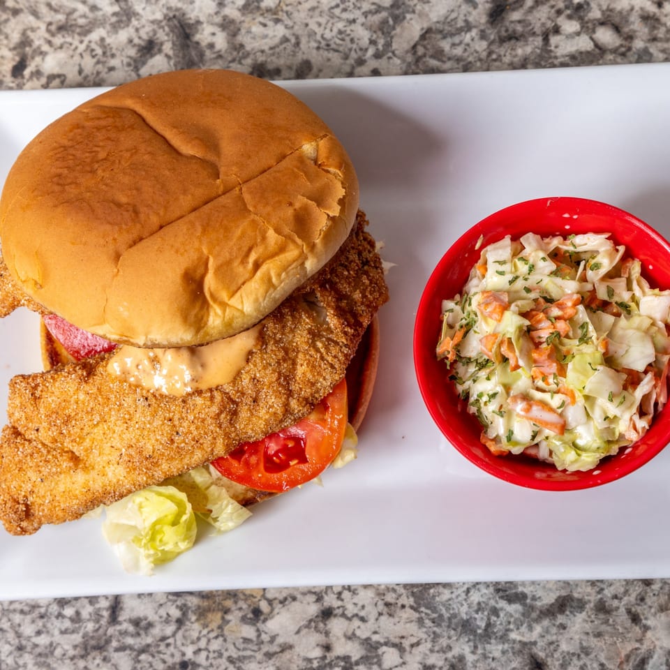 Crispy fried fish sandwich with lettuce, tomato, and sauce, served with a side of coleslaw.